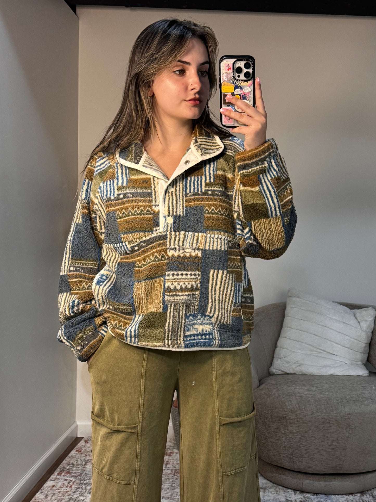 Person wearing a patterned shirt and green pants taking a mirror selfie in a room with a gray sofa and textured wall.