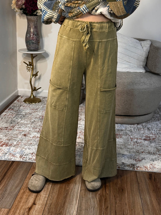 Person wearing a patterned top and green pants standing in a room with a couch and decor.