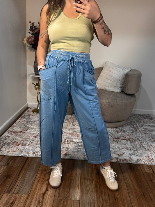 Person wearing a green top and blue barrel sweatpants standing in a room with a couch and wooden floor.