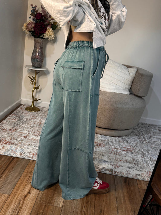 Person taking a mirror selfie wearing a light gray jacket and green pants in a room with a couch and wooden floor.