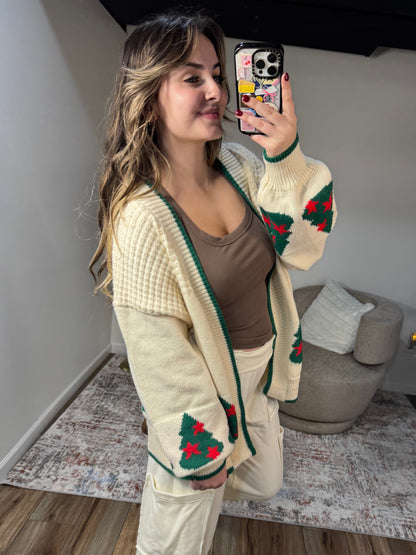 Person wearing a beige cardigan with red and green patterns, standing in a room with a couch and rug.