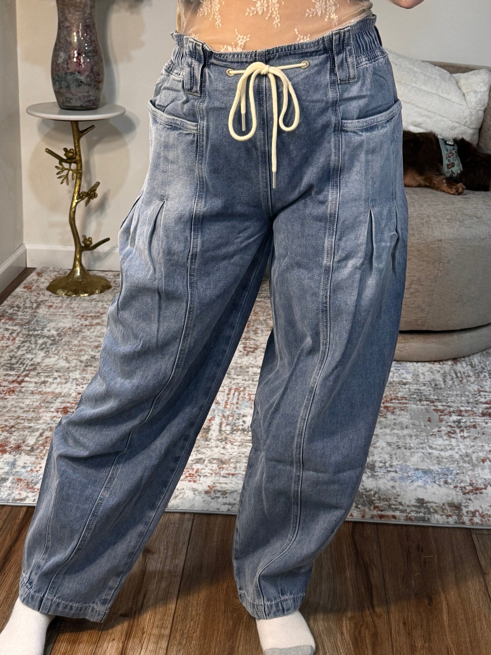 Person wearing blue jeans standing on a patterned rug with a couch and decorative items in the background.