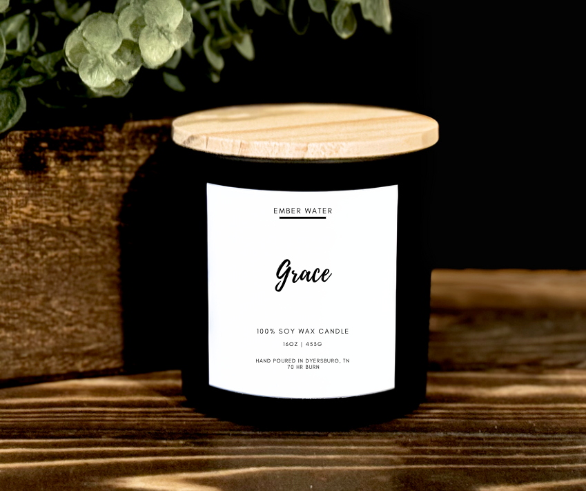 Candle labeled 'Grace' next to a potted plant on a wooden surface with a dark background