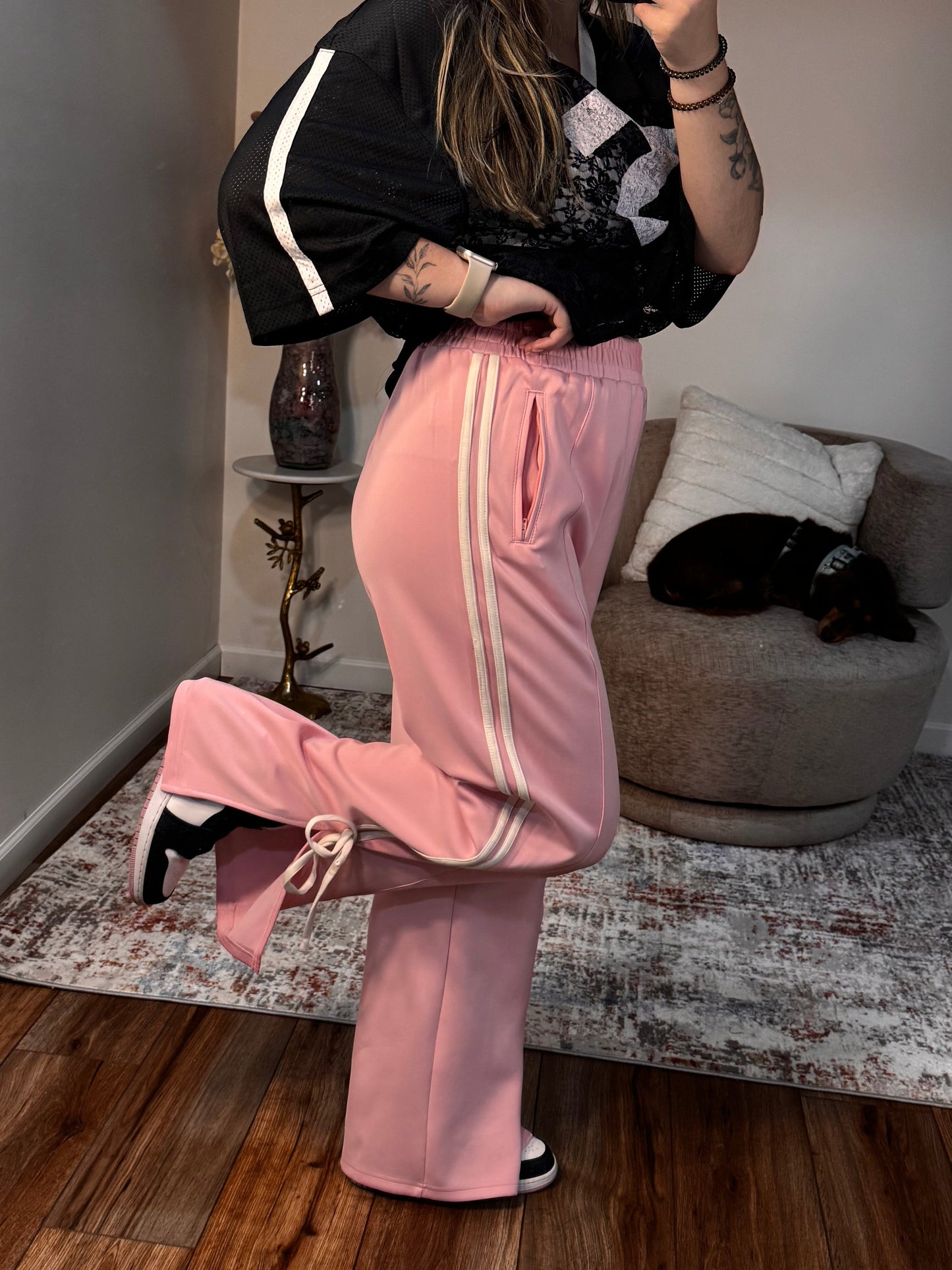 Person taking a mirror selfie wearing a black top and pink pants in a room with wooden flooring.