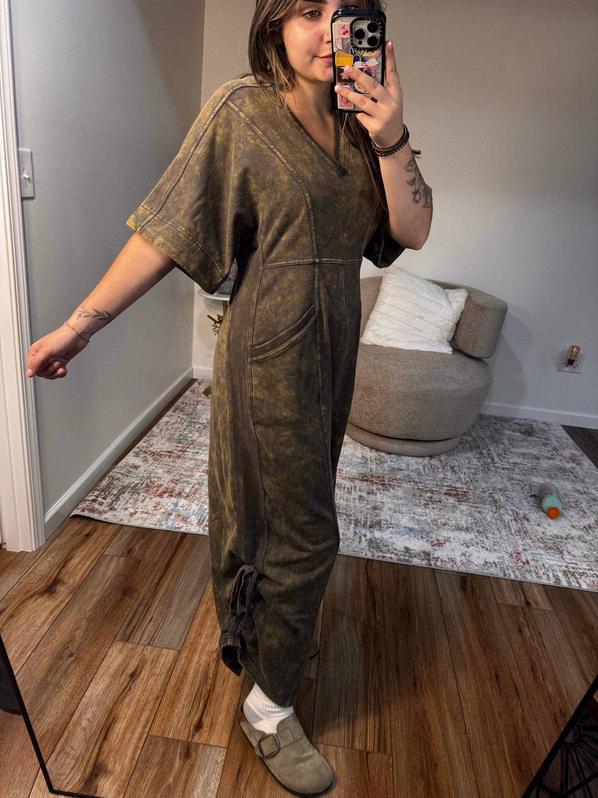 Person taking a mirror selfie wearing a brown jacket in a room with wooden flooring and a white wall.