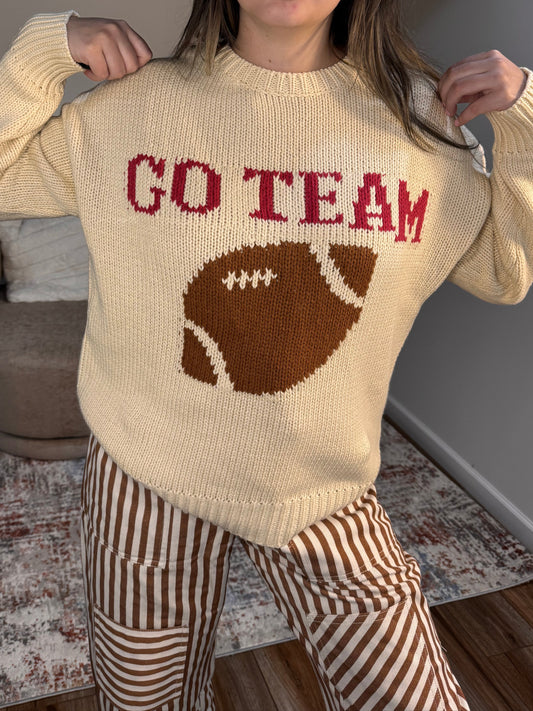 Person wearing a cream sweater with a football design and text indoors.