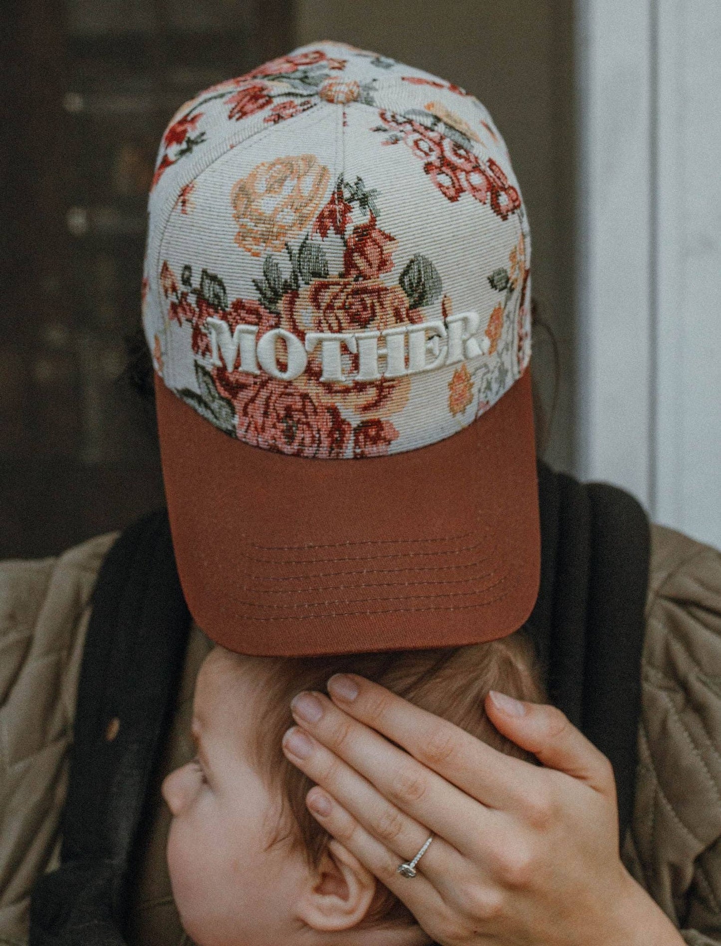 Person wearing a floral embroidered cap with 'MOTHER' text, holding a child.
