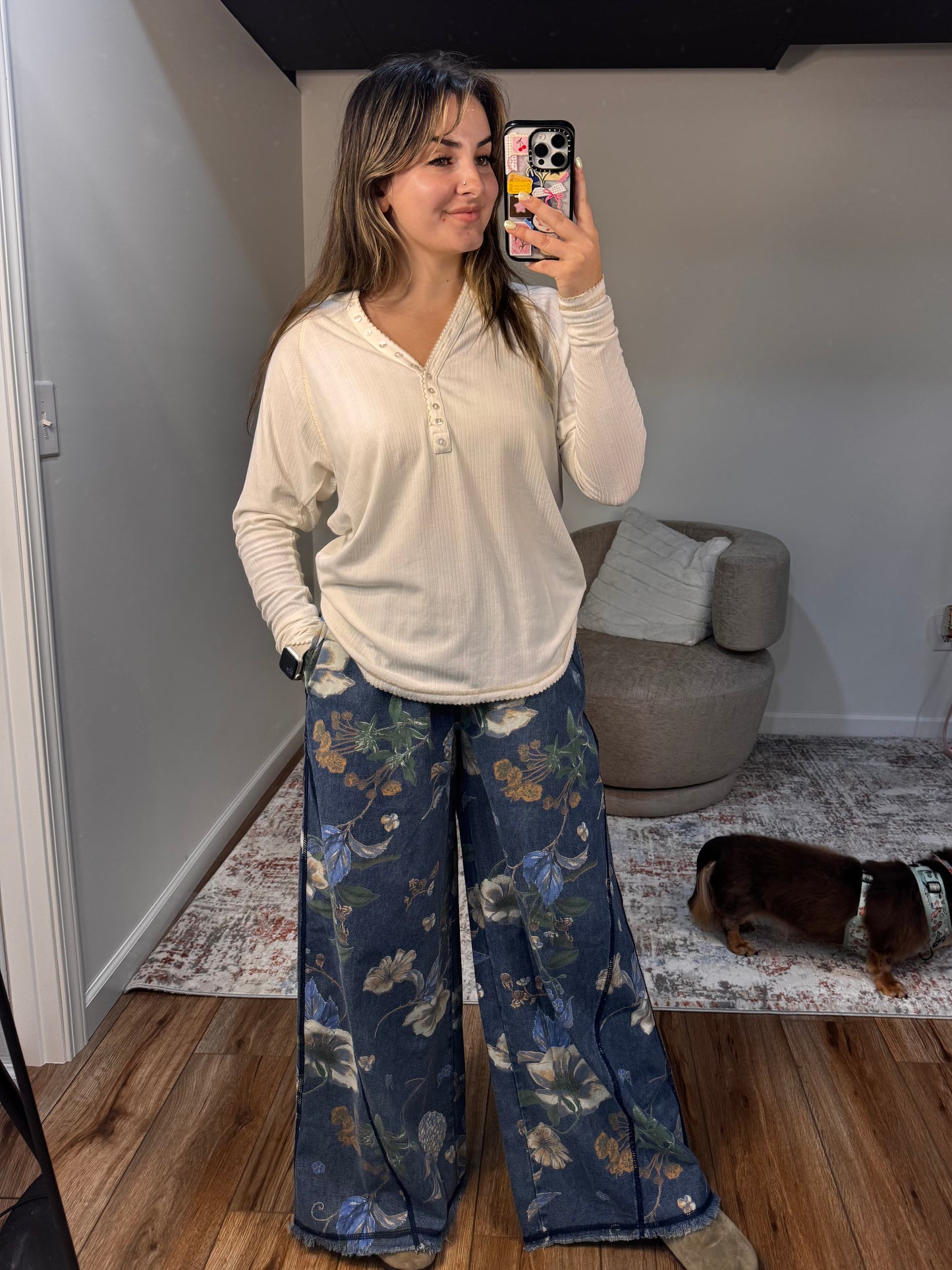 Woman taking a mirror selfie wearing a white long-sleeve shirt and blue floral pants in a room with a gray ottoman and rug.