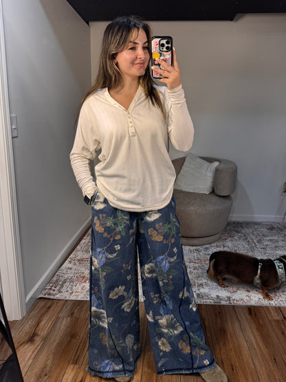 Woman taking a mirror selfie wearing a white long-sleeve shirt and blue floral pants in a room with a gray ottoman and rug.
