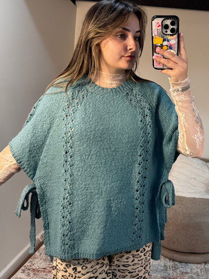 Woman taking a mirror selfie wearing a teal poncho in a living room.