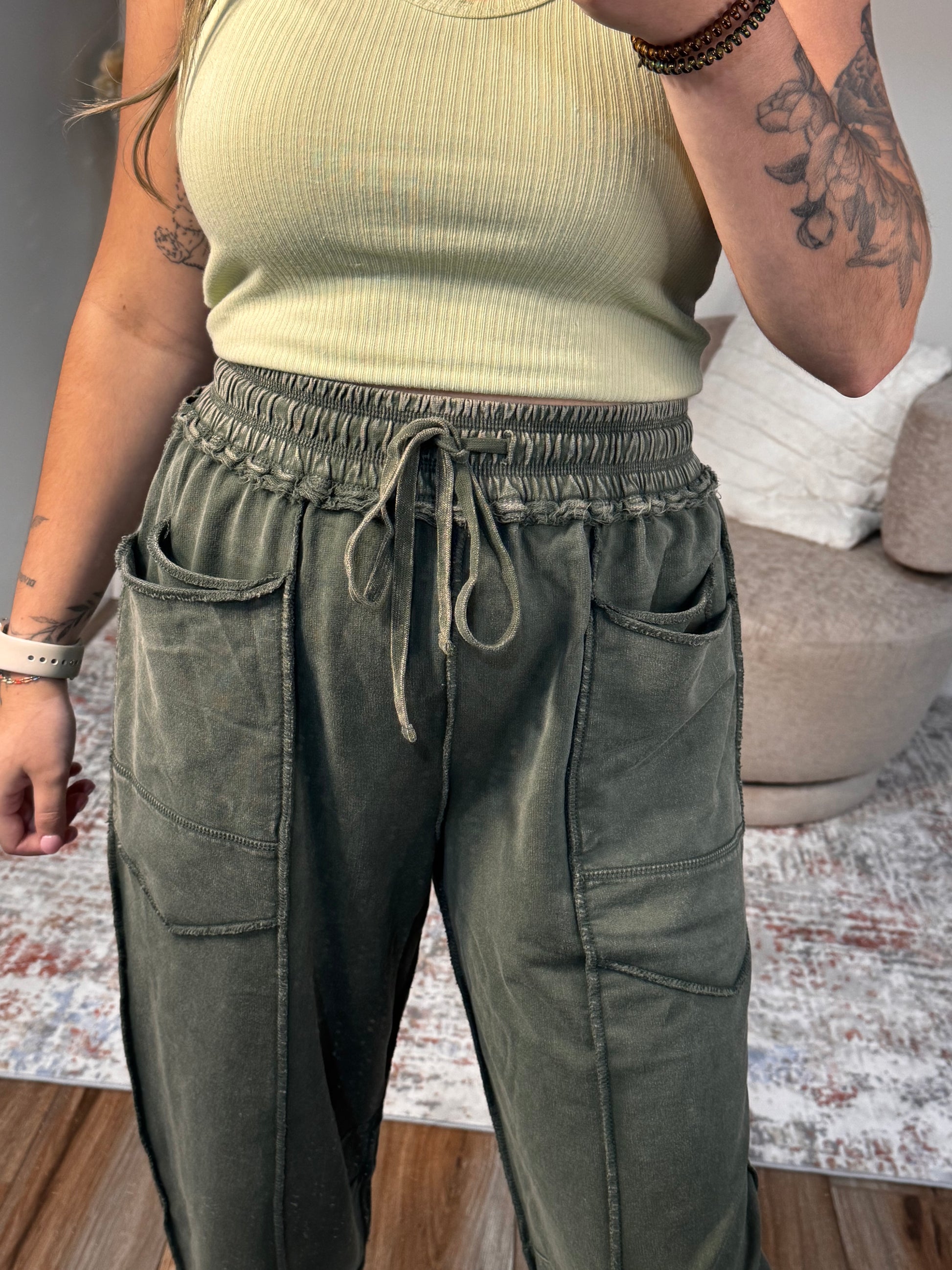Person wearing green pants and a beige top with tattoos visible on arms.