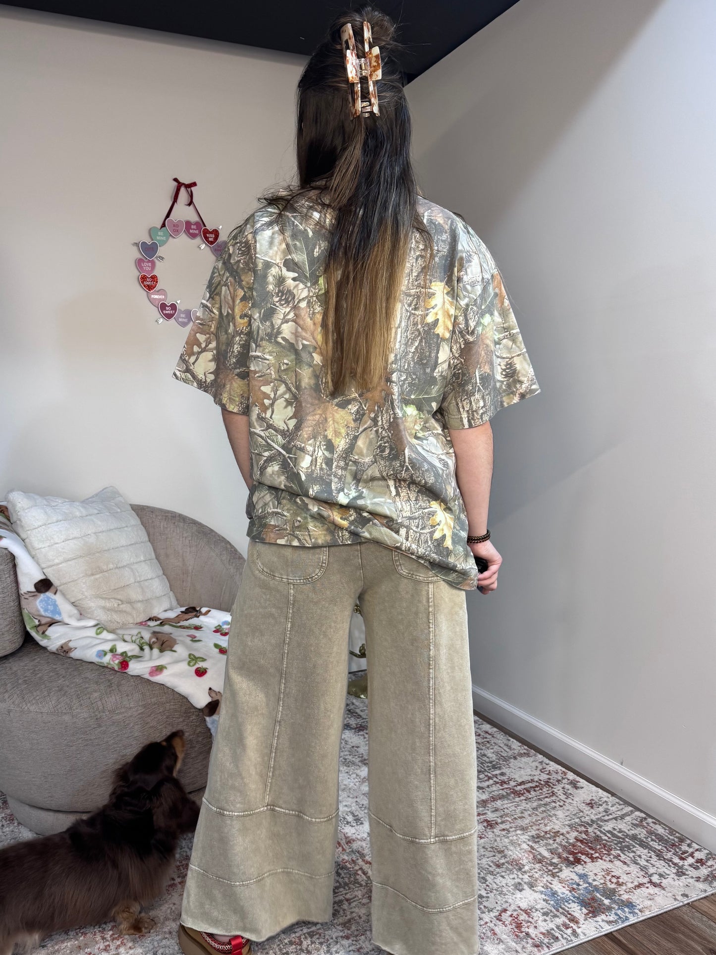 Person wearing a camouflage shirt and beige pants standing in a room with a couch and decorative items.