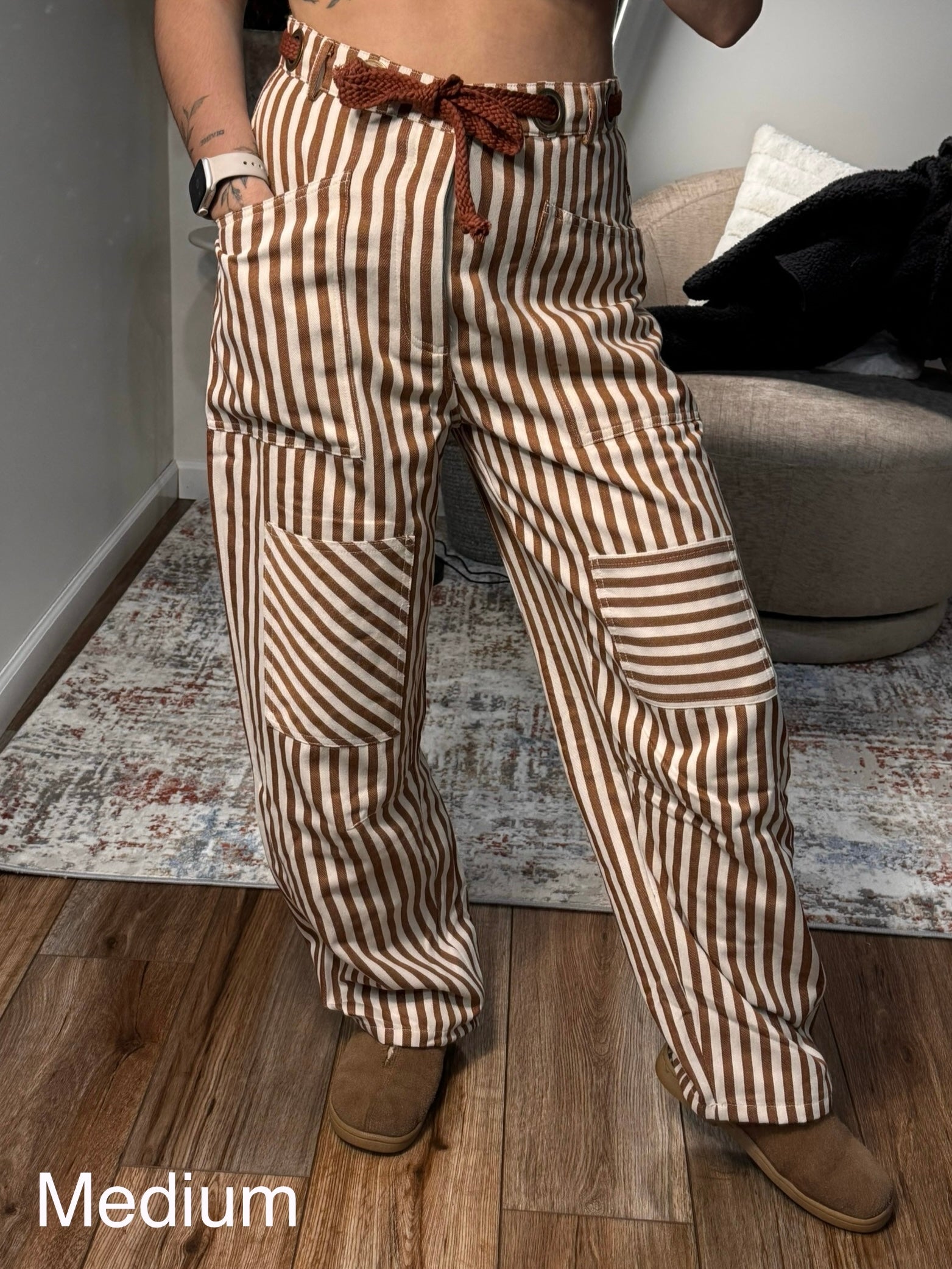 Person wearing brown and beige striped pants with a belt, standing in a room with a couch and rug.